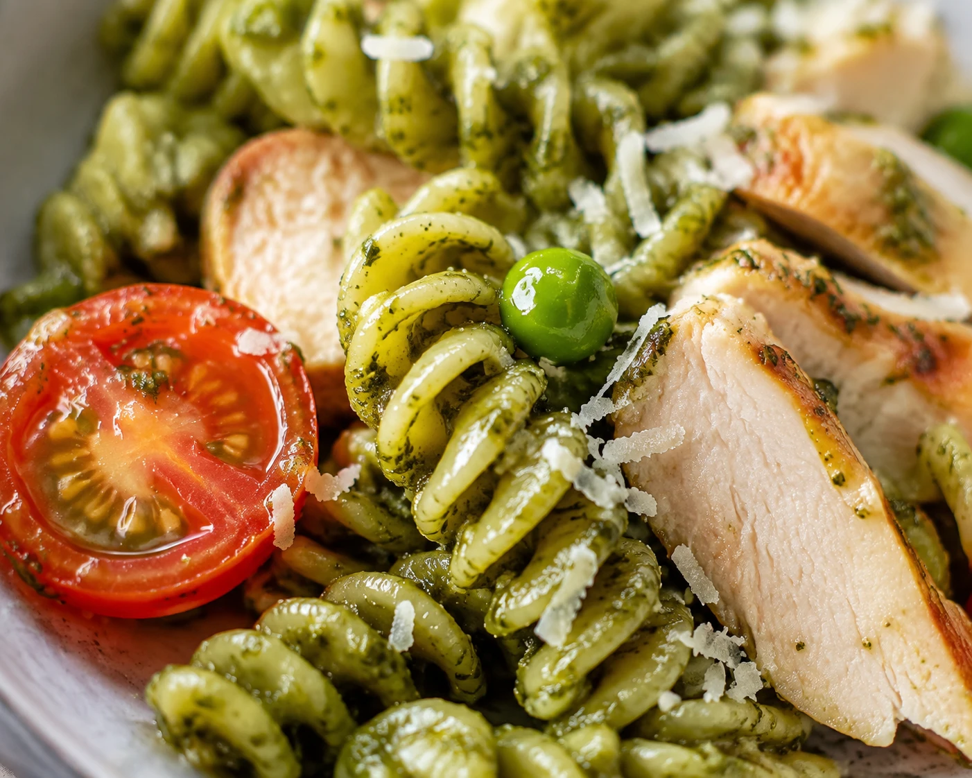 Glossy pesto rotini with a green pea, Parmesan, sliced chicken, and a halved cherry tomato in close detail.