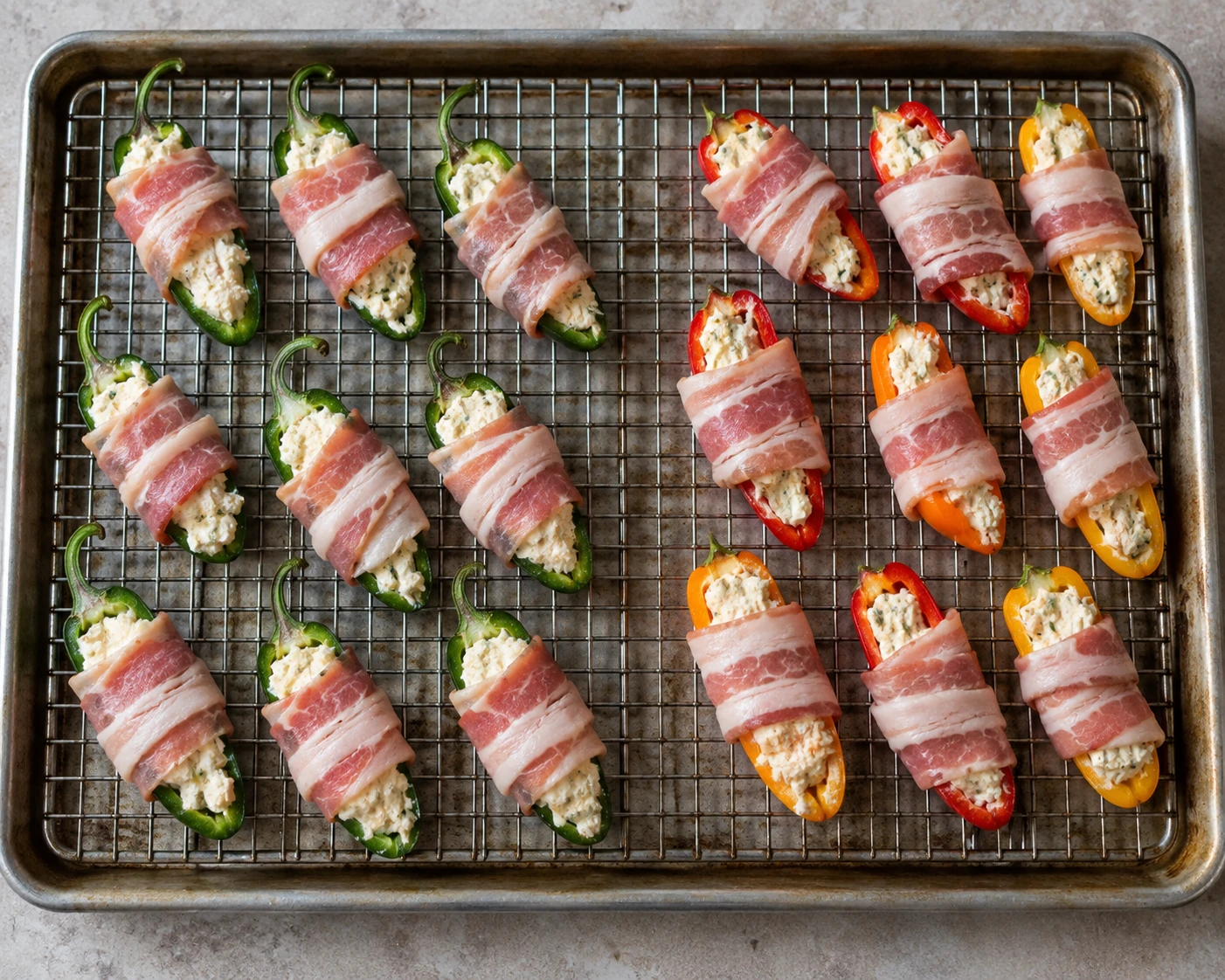 Filled jalapeño and sweet pepper halves are wrapped in bacon and arranged apart on a wire rack.