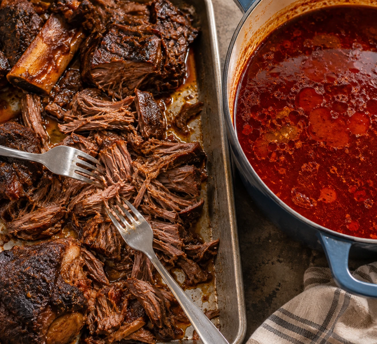 Tender braised beef is shredded beside a Dutch oven of chile-red birria broth and clarified fat.