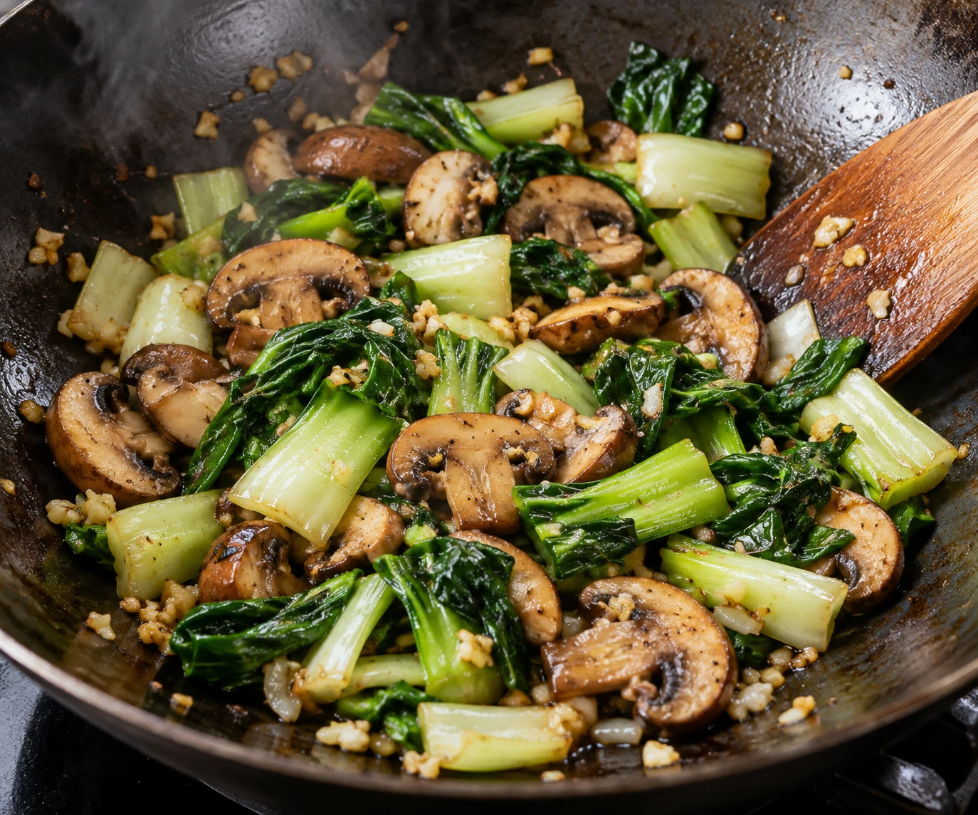 Mushrooms and bok choy stems and leaves stir-fry with garlic in a dark wok before noodles are added.