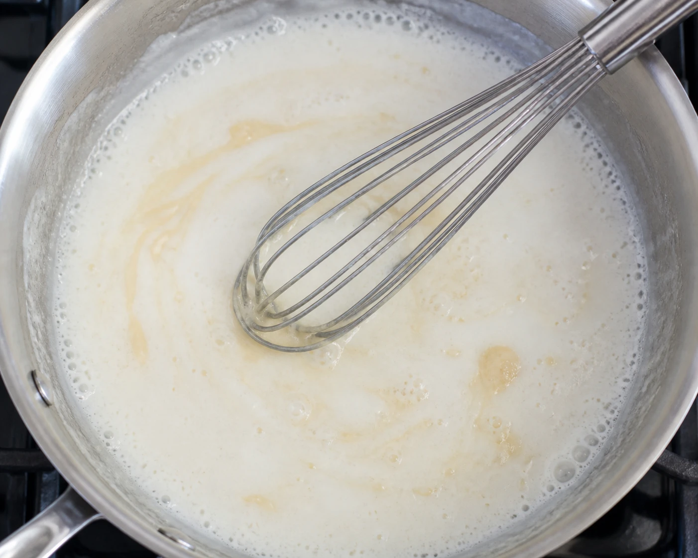 A whisk stirs milk into a pale roux in a skillet with gentle bubbles around the edge.