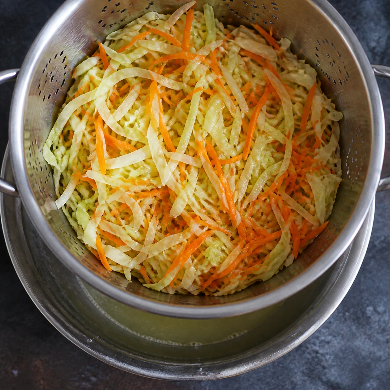 Salted shredded cabbage and carrots drain in a colander over a bowl before dressing.