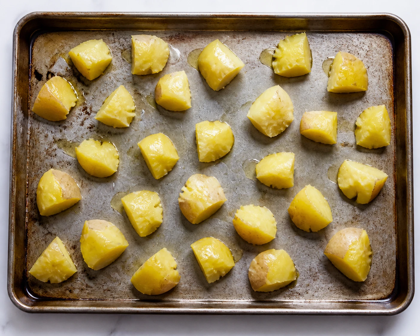 Roughened oil-coated Yukon Gold potato chunks are spaced across a rimmed sheet pan before roasting.