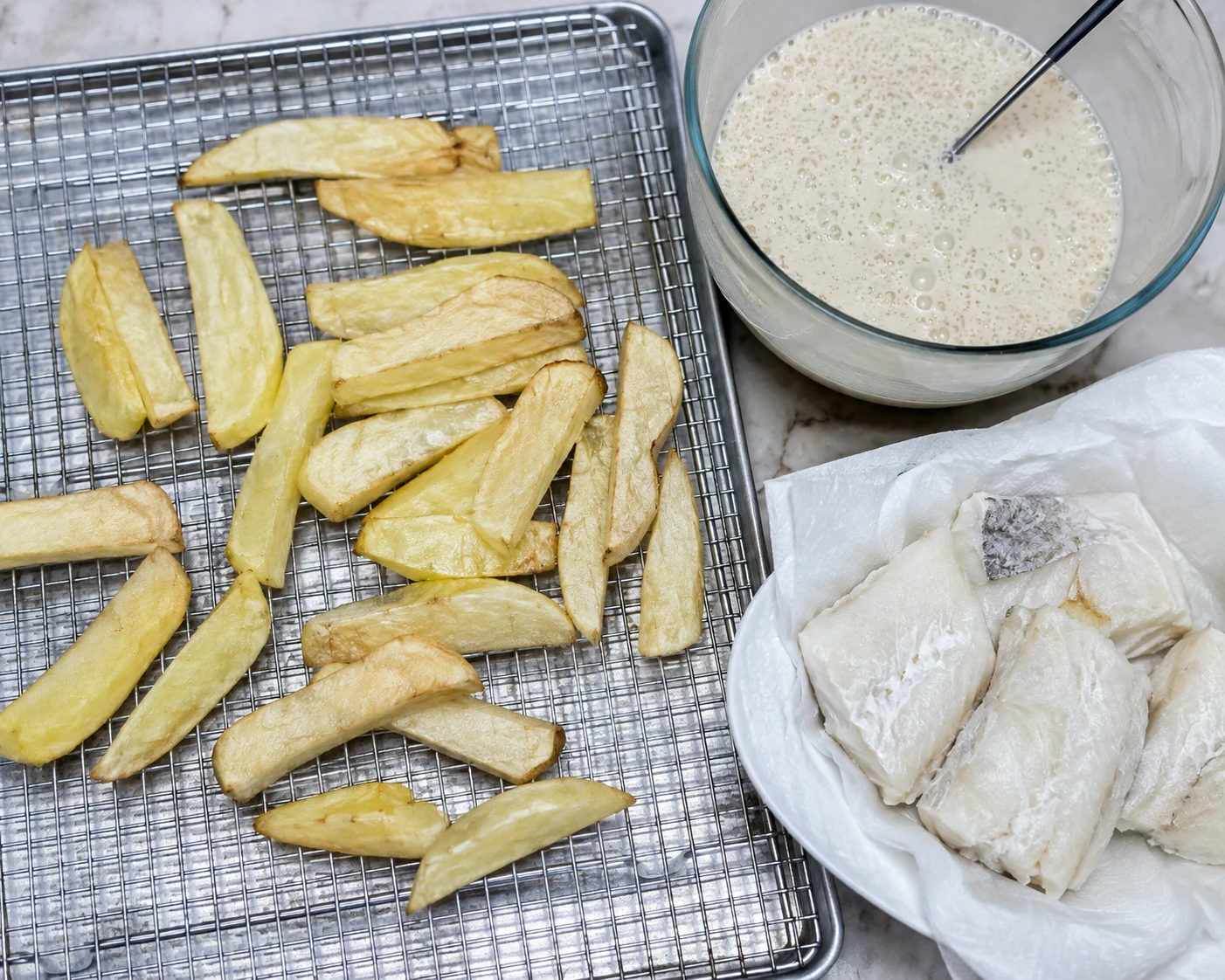 Pale first-fried chips drain on a rack beside batter and prepared cod portions.