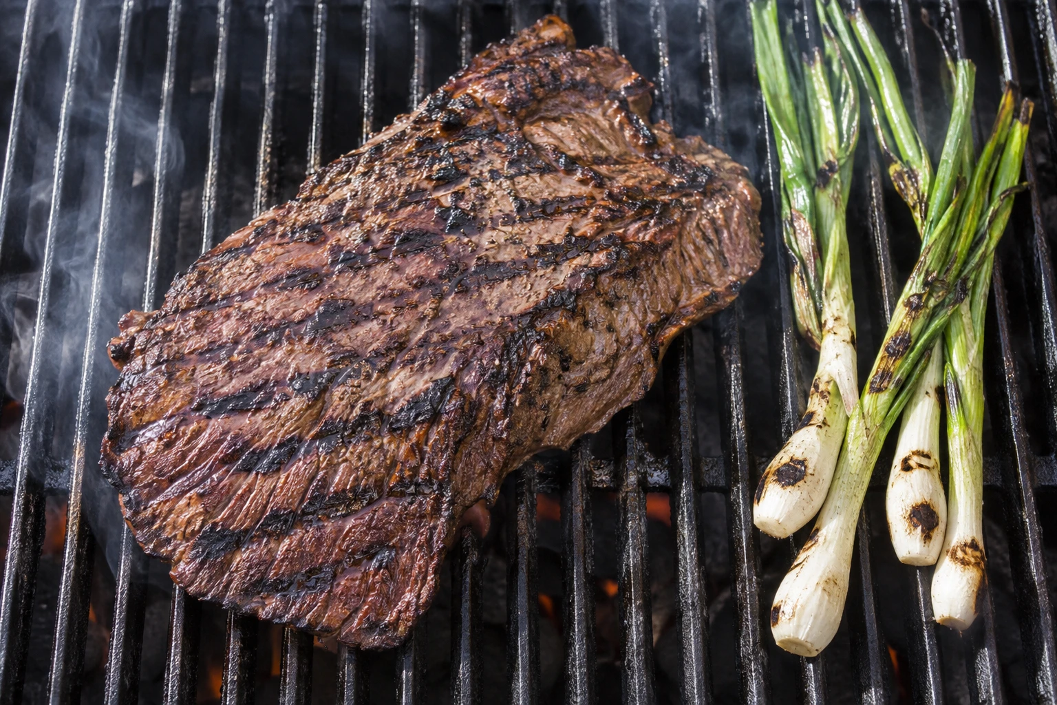 A whole flank steak sears over hot grill grates while scallions char alongside it.