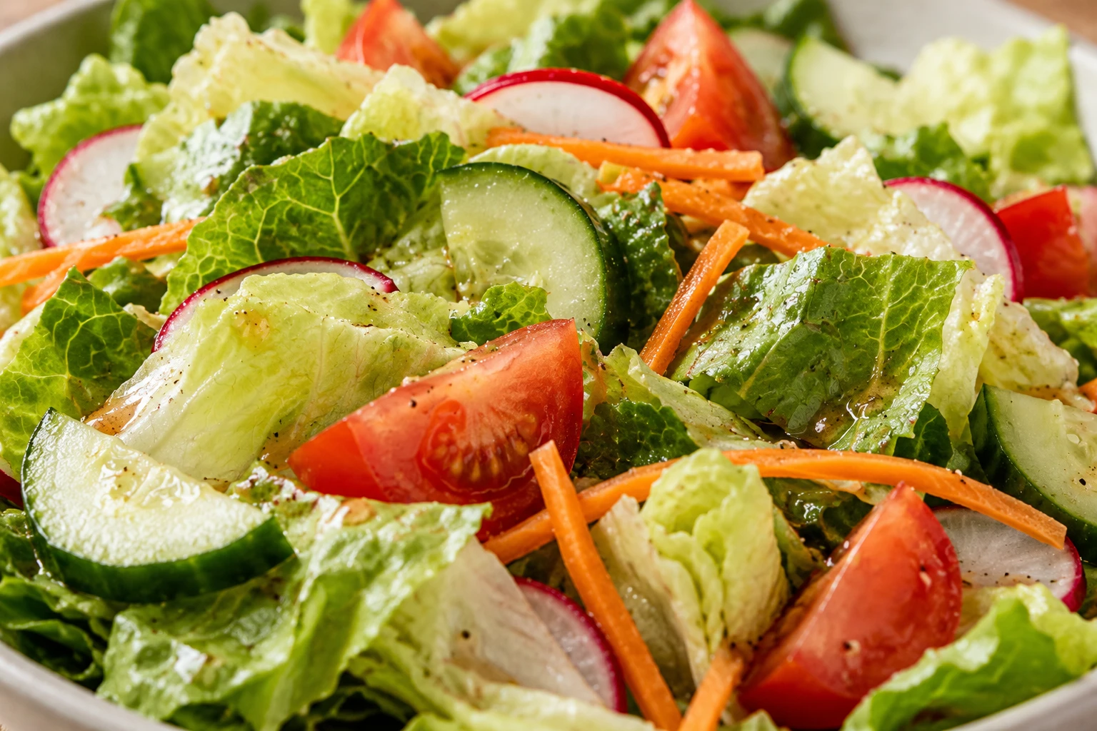 Light vinaigrette clings to crisp lettuce beside distinct cucumber, tomato, carrot, and radish.
