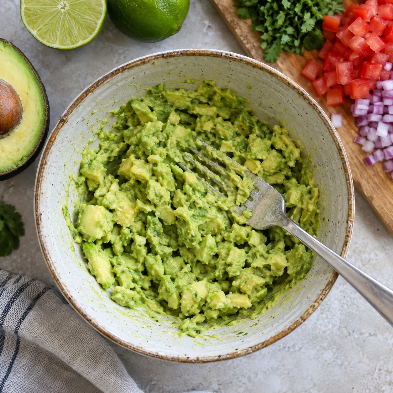 A fork mashes lime-seasoned avocado into a chunky base in a ceramic bowl before vegetables are folded in.