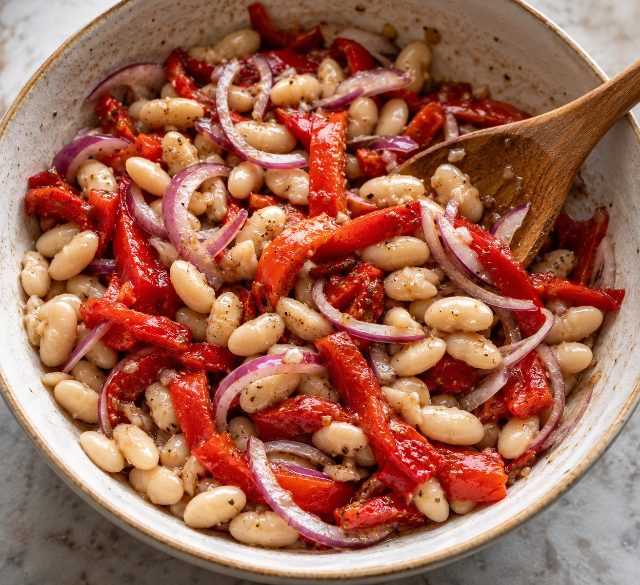 A wide spoon gently folds white beans, red pepper strips, and red onion through vinaigrette.