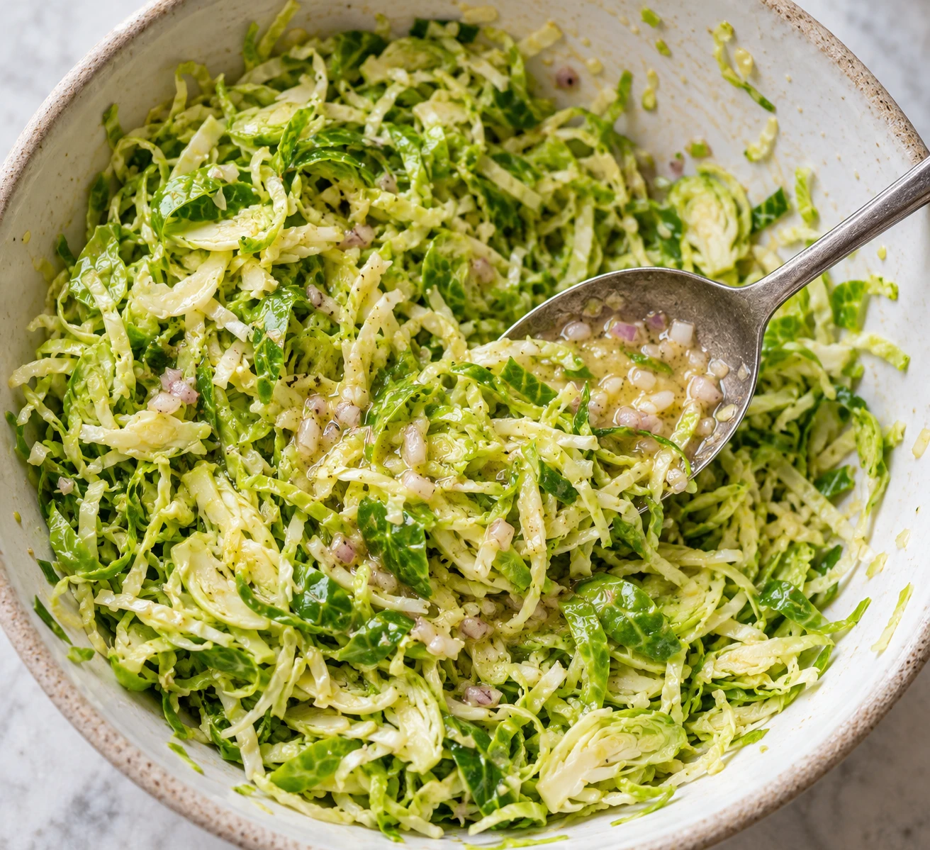 Fine raw Brussels sprout ribbons receive a light coating of lemon-shallot vinaigrette in a large bowl.