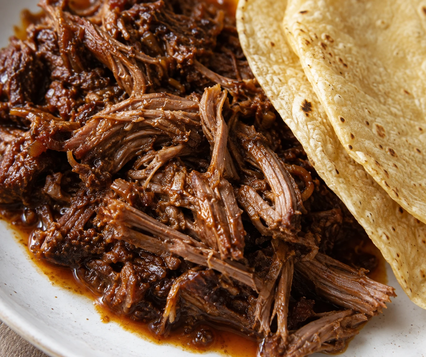 Tender glossy strands of beef barbacoa sit beside a folded warm corn tortilla.