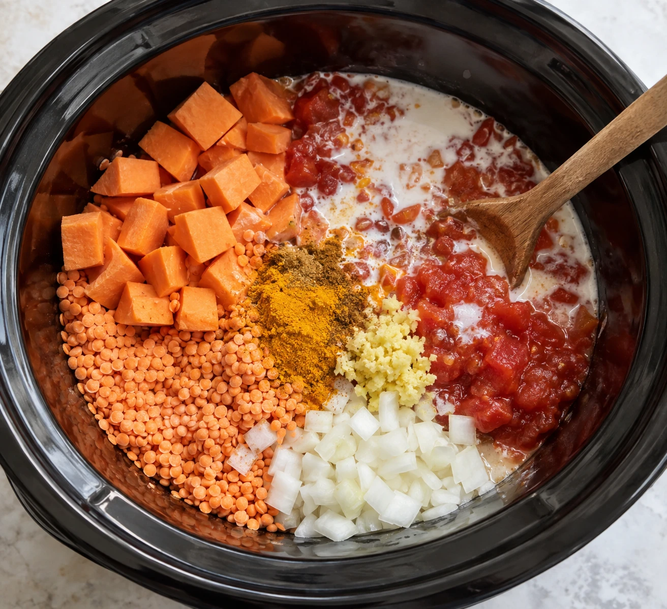 A slow cooker holds red lentils, sweet potato, tomatoes, coconut milk, onion, ginger, and spices before cooking.