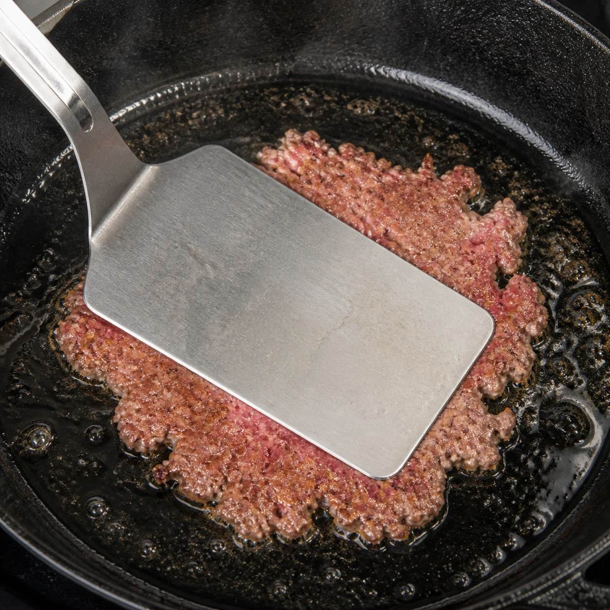 A metal spatula presses a loose beef ball into a thin smash-burger patty on a cast-iron skillet.
