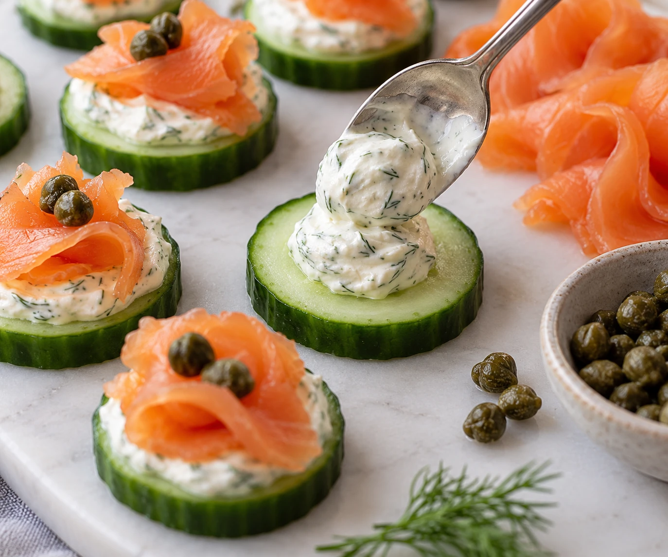 A small spoon places dill crème fraîche on a dry cucumber round beside salmon and capers.