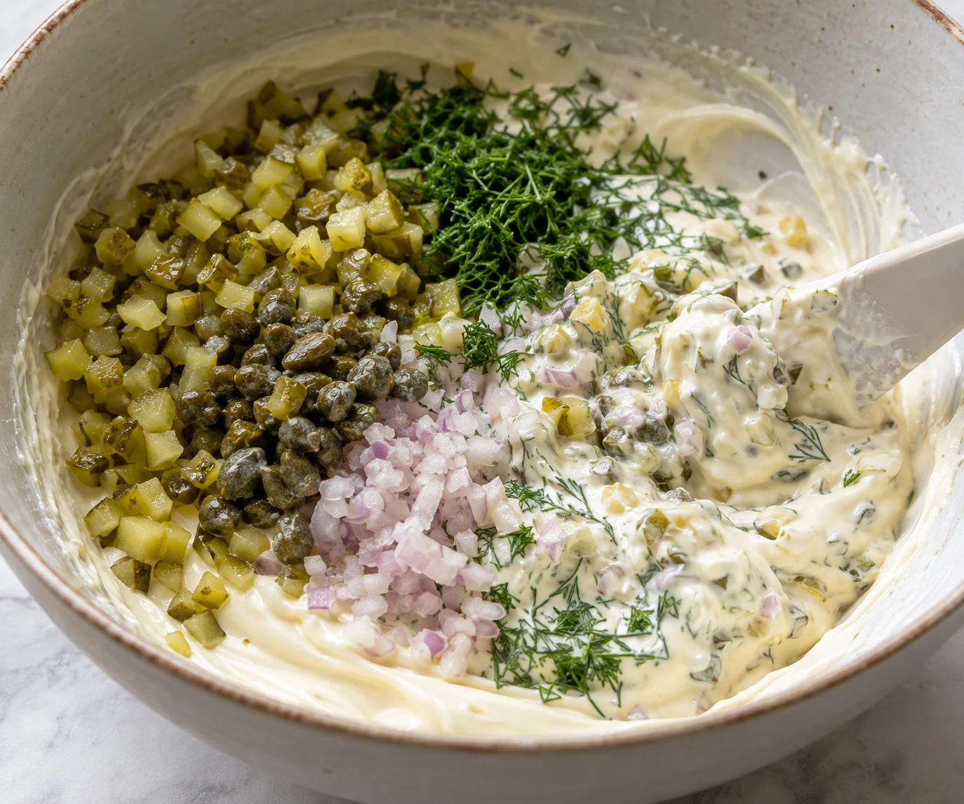 Finely chopped pickles, capers, shallot, and dill are being folded into mayonnaise.
