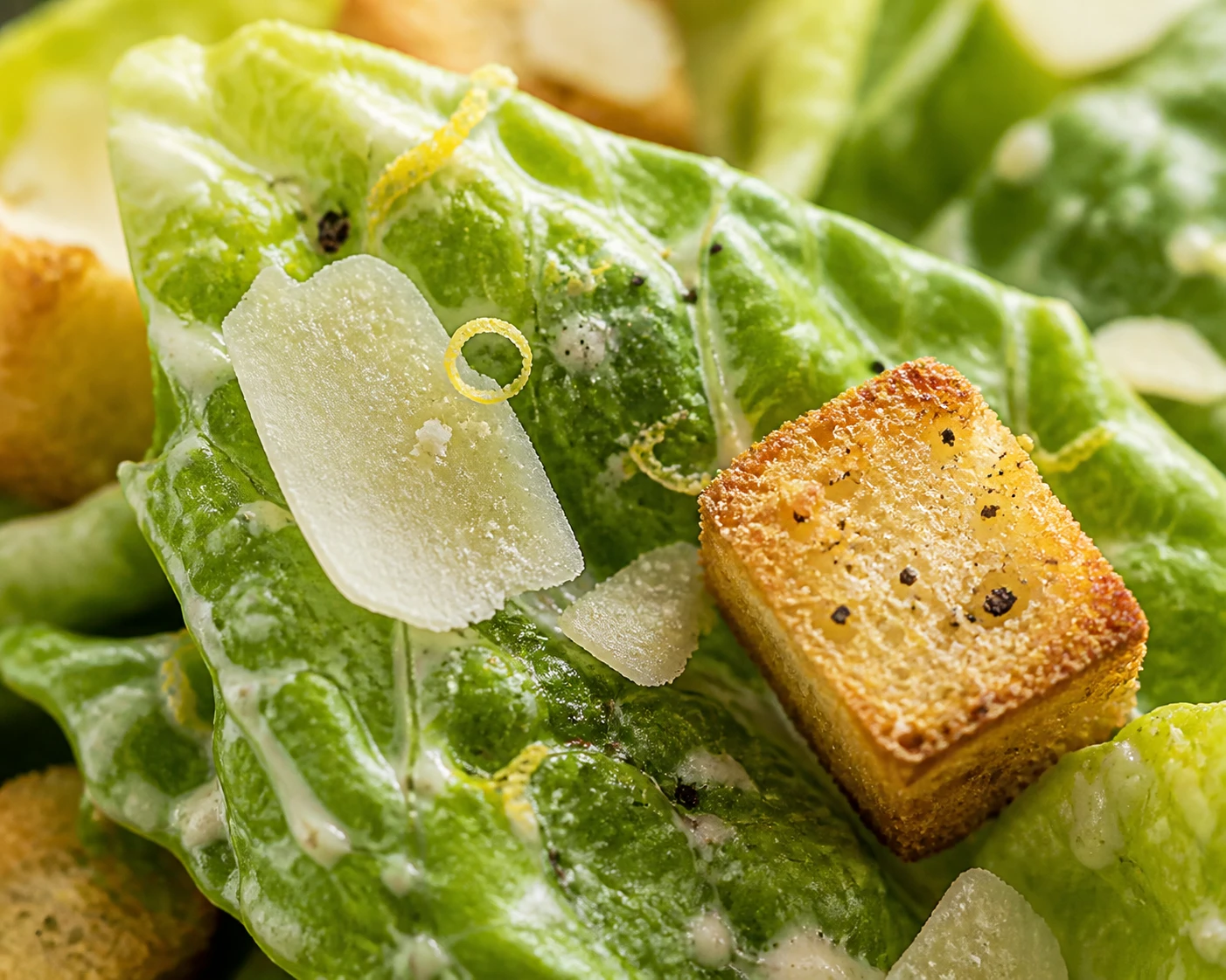 A close Caesar salad bite shows creamy romaine, shaved Parmesan, lemon zest, toasted pepper, and a crisp crouton.